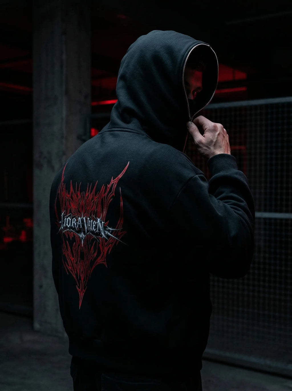 Person wearing a black hoodie with a red and white logo on the back, standing in a dark setting.